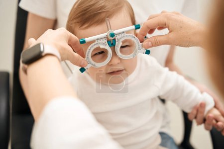 Téléchargez les photos : Mains optométristes portant le cadre d'un essai ophtalmique sur un petit patient tranquille en présence de sa mère - en image libre de droit