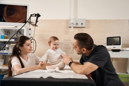 Téléchargez les photos : Endoscopiste sympathique communiquant avec l'enfant en présence de sa jeune mère heureuse dans la salle d'opération - en image libre de droit