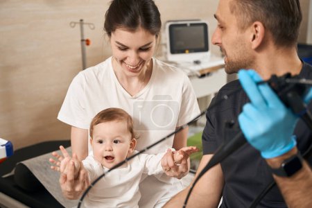 Téléchargez les photos : Jeune médecin avec endoscope à la main regardant la mère souriante et son bébé - en image libre de droit