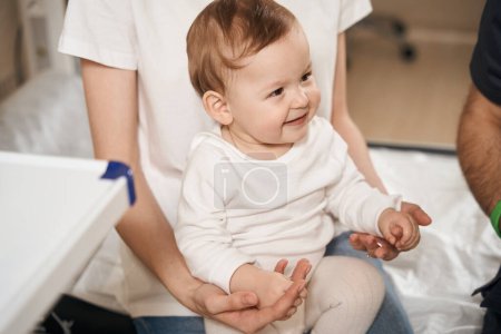 Téléchargez les photos : Petit enfant agréable assis sur les genoux du parent féminin à côté du médecin masculin - en image libre de droit