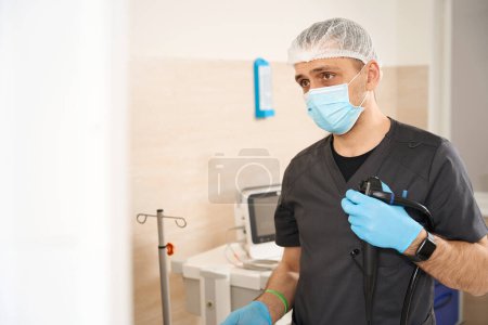 Waist-up portrait of endoscopist in face mask and disposable gloves holding endoscope