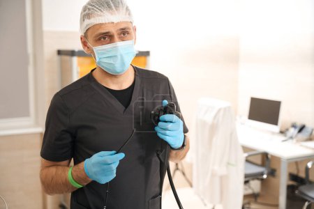 Téléchargez les photos : Portrait taille haute de l'endoscopiste en masque médical et gants jetables tenant l'endoscope avec tube d'insertion - en image libre de droit