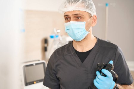 Foto de Retrato de endoscopista serio en máscara facial desechable que sostiene el endoscopio con la mano enguantada - Imagen libre de derechos