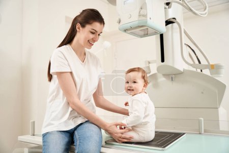 Foto de Joven alegre poniendo a su hijo pequeño en la mesa de rayos X en la clínica - Imagen libre de derechos