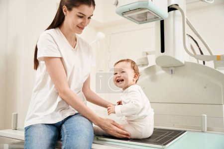 Téléchargez les photos : Agréable jeune parent féminin plaçant son bébé sur la table radiographique dans une clinique privée - en image libre de droit