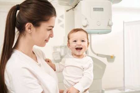 Foto de Mujer sonriente sosteniendo a su niño alegre frente a la máquina digital de rayos X - Imagen libre de derechos