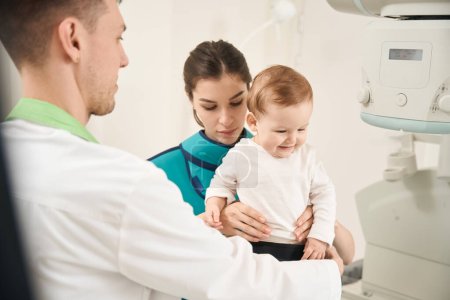 Photo for Radiographer putting protective clothing on infant in presence of serious female parent - Royalty Free Image