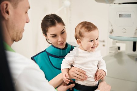 Téléchargez les photos : Radiologue mettre des vêtements de protection sur bébé heureux en présence d'une jeune mère souriante - en image libre de droit