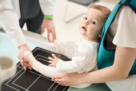 Foto de Foto recortada del radiólogo que coloca la pierna del niño en la mesa radiográfica ayudada por el padre femenino - Imagen libre de derechos