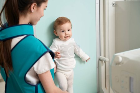 Téléchargez les photos : Jeune femme habillée en tablier de plomb mettant bébé contre le mur devant la machine à rayons X - en image libre de droit