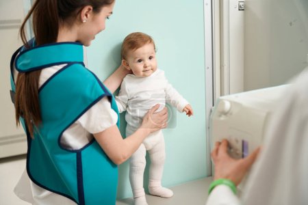 Foto de Calma bebé de pie por la pared delante de la máquina de rayos X con el apoyo de la madre cariñosa - Imagen libre de derechos