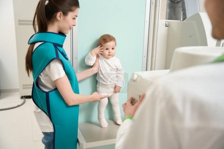 Photo for Peaceful child standing against wall in front of x-ray machine supported by young mother in lead apron - Royalty Free Image