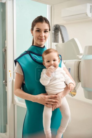 Téléchargez les photos : Femme souriante dans le tablier de plomb tenant bébé dans ses bras tout en se tenant à côté de l'appareil à rayons X - en image libre de droit