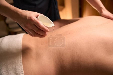 Photo for Cropped photo of qualified masseuse pouring organic oil from ceramic bowl on naked back of client - Royalty Free Image