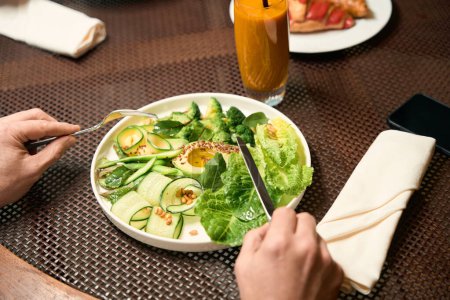 Téléchargez les photos : Photo recadrée de mains masculines tenant couteau et fourchette sur une assiette en céramique avec salade de légumes frais - en image libre de droit