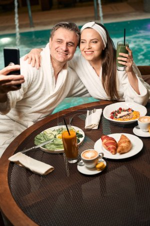 Téléchargez les photos : Femme romantique étreignant son compagnon heureux à la table du déjeuner pendant la séance photo près de la piscine - en image libre de droit