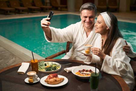 Photo for Man in love embracing his contented female companion during photo shoot at lunch table by swimming pool - Royalty Free Image