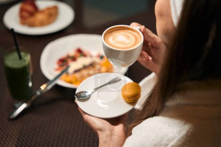 Photo for Cropped photo of female in bathrobe sitting at breakfast table with cup of coffee in her hand - Royalty Free Image