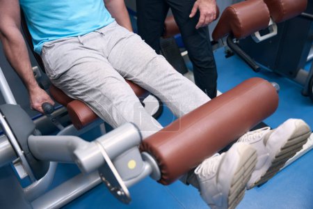 Téléchargez les photos : Photo recadrée d'une personne en forme faisant une extension de jambe assise sur une machine de musculation en présence d'un instructeur de gym - en image libre de droit
