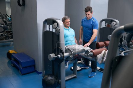 Téléchargez les photos : Homme athlétique agréable effectuant l'extension des jambes assises sur une machine d'exercice supervisée par un entraîneur de fitness - en image libre de droit