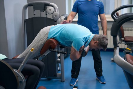 Téléchargez les photos : Homme adulte effectuant une extension du dos sur une machine d'exercice supervisée par un entraîneur personnel - en image libre de droit