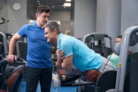 Téléchargez les photos : Homme adulte joyeux faisant de l'exercice sur banc d'hyperextension sous la direction d'un entraîneur de gym - en image libre de droit