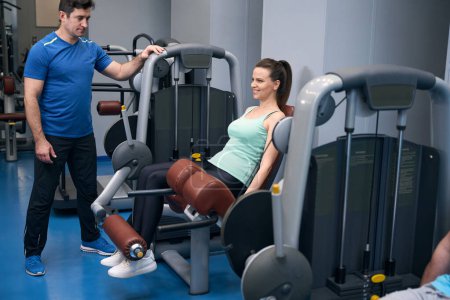 Photo for Cheerful mature lady performing seated leg extension on exercise machine under guidance of gym instructor - Royalty Free Image