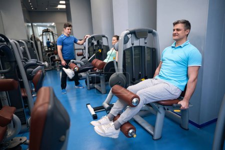 Téléchargez les photos : Femme et homme souriants effectuant une extension de jambe assise sur un équipement de gymnastique sous la direction d'un entraîneur - en image libre de droit