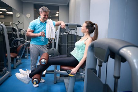 Téléchargez les photos : Femme souriante exerçant sur la machine d'extension de jambe tout en parlant à compagnon d'entraînement heureux - en image libre de droit