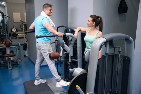 Téléchargez les photos : Homme sportif agréable effectuant élévation des jambes latérales sur la machine d'exercice en présence d'un partenaire de fitness souriant - en image libre de droit