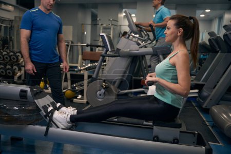 Photo for Smiling fit woman exercising on seated row machine under supervision of fitness instructor - Royalty Free Image