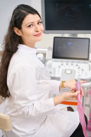 Photo for Smiling female cardiologist in white coat sitting near equipment and holding device in the hospital - Royalty Free Image