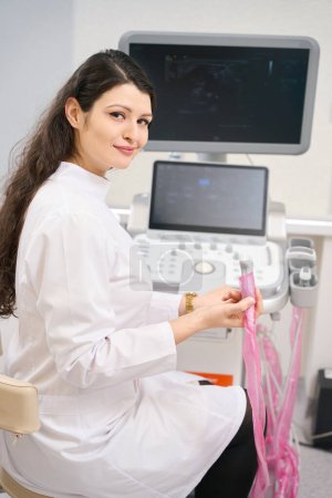 Photo for Young doctor in white coat sitting near equipment and holding device, looking at the camera - Royalty Free Image