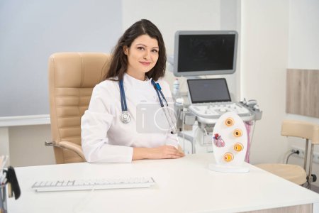 Téléchargez les photos : Cardiologue féminine en manteau blanc avec stéthoscope assis à table et regardant la caméra - en image libre de droit