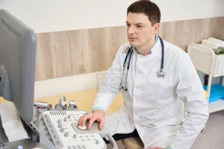 Téléchargez les photos : Médecin en manteau blanc assis près de l'équipement et regardant le moniteur à l'hôpital - en image libre de droit