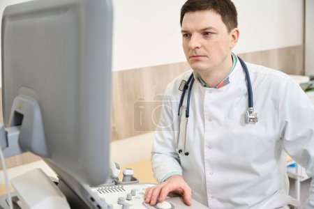 Téléchargez les photos : Cardiologue en blouse blanche assis près de l'échographie et regardant le moniteur dans la clinique - en image libre de droit