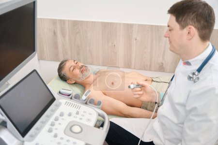 Doctor in white coat sitting near ultrasound machine and holding scanner, looking at patient