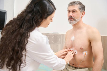 Téléchargez les photos : Infirmière en manteau blanc préparant le patient à l'électrocardiographie du cœur, homme regardant le médecin - en image libre de droit