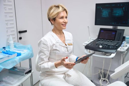 Téléchargez les photos : Amical gynécologue femme souriant tout en étant assis dans le bureau avec une tablette dans ses mains - en image libre de droit