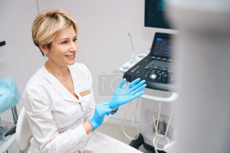 Foto de Ginecóloga sonriente que usa guantes médicos antes de examinar a la paciente. Foto de una agradable doctora en proceso de trabajo - Imagen libre de derechos