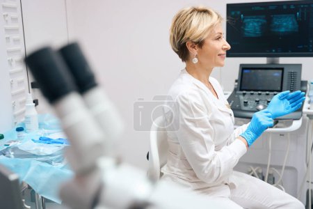 Foto de Foto de una ginecóloga amable que se pone guantes médicos antes de examinar al paciente con el fondo de la máquina de ultrasonido. - Imagen libre de derechos