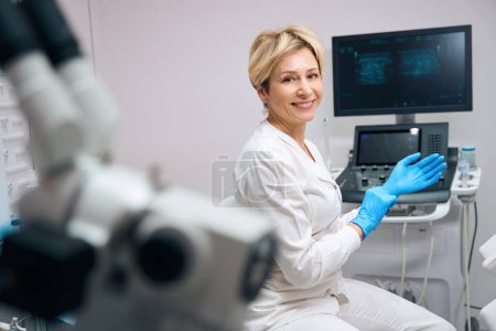 Téléchargez les photos : Portrait de femme souriante au gynécologue qui porte des gants médicaux jetables avant d'examiner le patient sur fond de machine à échographie - en image libre de droit