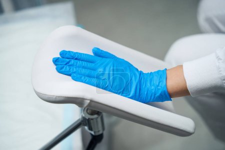 Téléchargez les photos : Photographie d'une main dans un gant médical a été placé sur la chaise gynécologique par le médecin - en image libre de droit