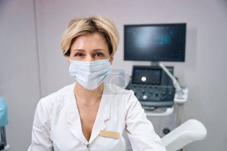 Foto de Foto de una ginecóloga que trabaja en una máscara médica contra el fondo de una máquina de ultrasonido - Imagen libre de derechos