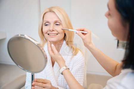Foto de Mujer bonita se somete a un procedimiento de limpieza facial ultrasónico. Señora se mira en el espejo mientras está sentado en la oficina esteticista - Imagen libre de derechos
