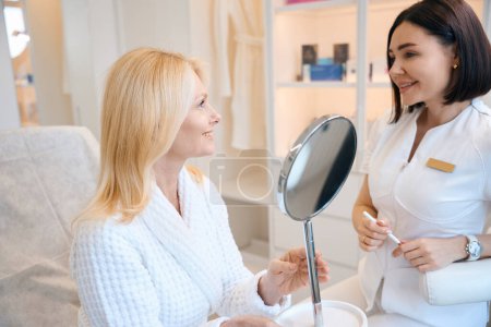 Téléchargez les photos : Femme joyeuse dans un centre de cosmétologie parle avec un cosmétologue après une procédure de nettoyage du visage par ultrasons - en image libre de droit