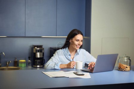 Téléchargez les photos : Joyeux employé de bureau lisant quelque chose sur l'écran d'ordinateur portable tout en étant assis au comptoir du bar dans la cuisine d'entreprise - en image libre de droit