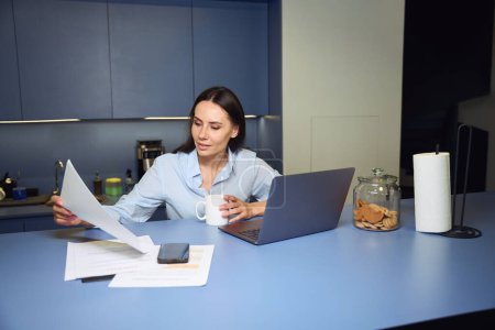 Téléchargez les photos : Employée regardant le document dans sa main alors qu'elle était assise au comptoir du bar dans la cuisine d'entreprise - en image libre de droit
