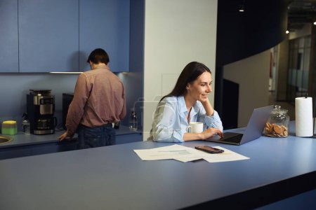 Téléchargez les photos : Travailleur d'entreprise souriant travaillant sur son ordinateur portable tandis que son collègue se faire du café - en image libre de droit