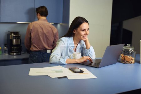 Téléchargez les photos : Joyeuse employée assise à table lisant quelque chose sur un écran d'ordinateur portable pendant que son collègue se faisait du café - en image libre de droit